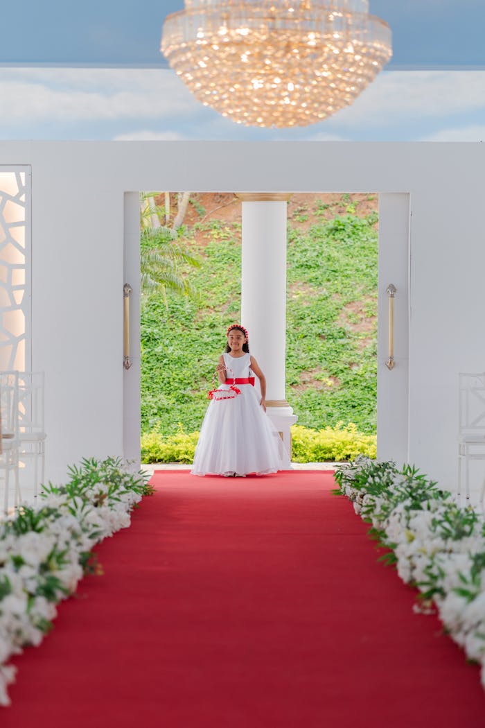 A young flower girl stands adorably at a wedding aisle decorated with white flowers and a red carpet.